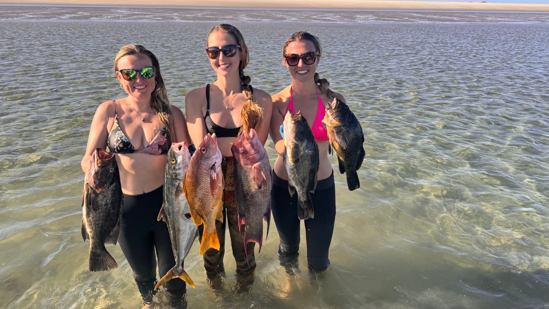 Three people standing in shallow water holding various fish.