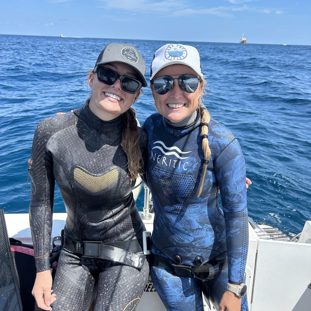 Two people in wetsuits and hats on a boat with ocean in the background.