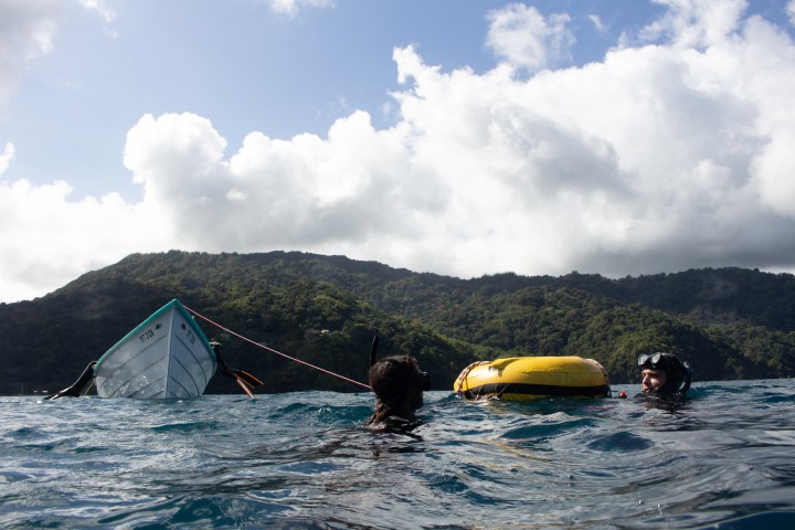 Two divers in water near a partially submerged boat, with a forested hill in the background.