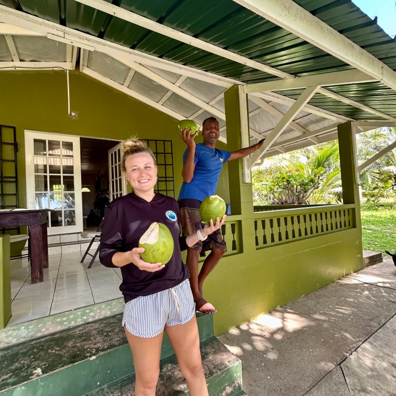 Two people smiling, holding coconuts on a porch of a green house.