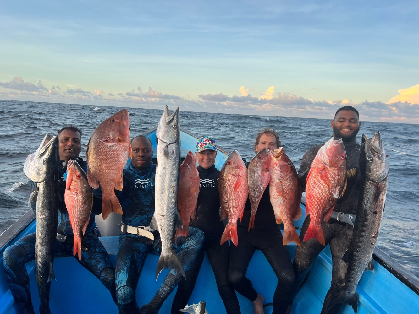 Five people in wetsuits on a boat holding large fish.