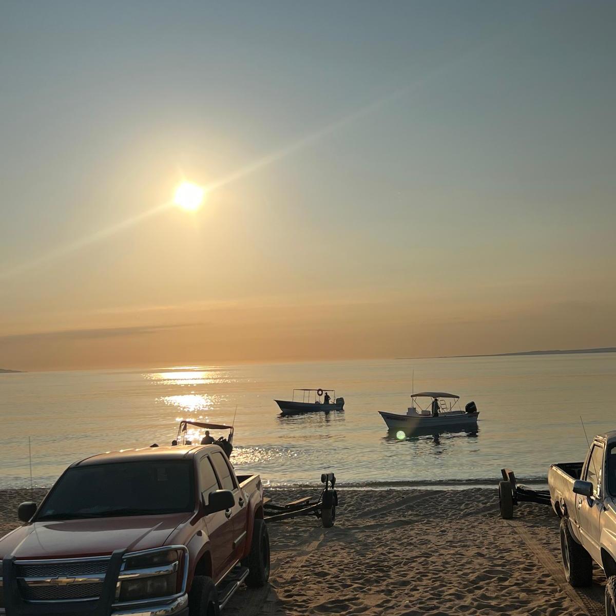 Sunset over beach with two boats on water and parked trucks on sand