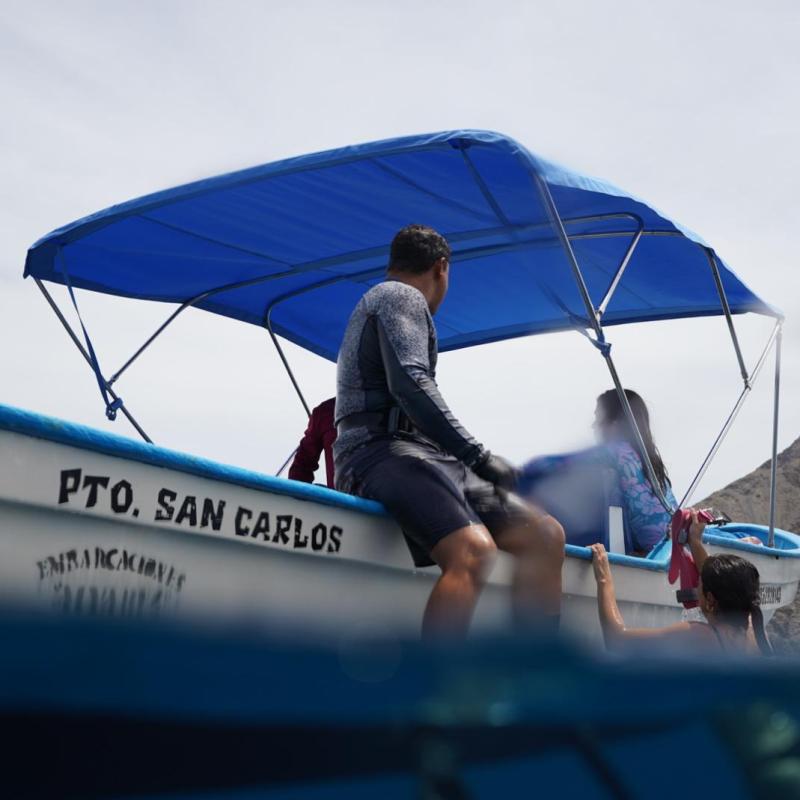 People sitting on a boat with a blue canopy near a mountain.