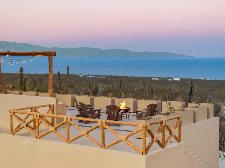 Rooftop patio with chairs around a fire pit overlooking the ocean and hills at sunset.